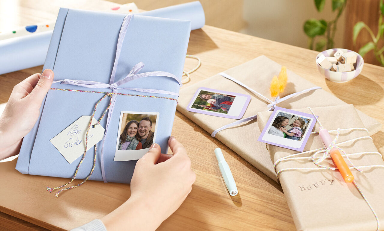 Una persona tiene un regalo impacchettato in modo creativo con foto Polaroid sopra. Più regali adorabilmente impacchettati sul tavolo.