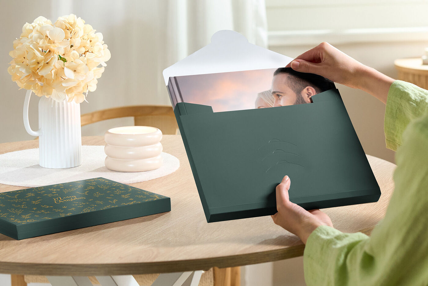 Una persona apre una scatola verde con un album fotografico su un tavolo. All'interno c'è un'immagine bellissima, circondata da fiori e design creativo.