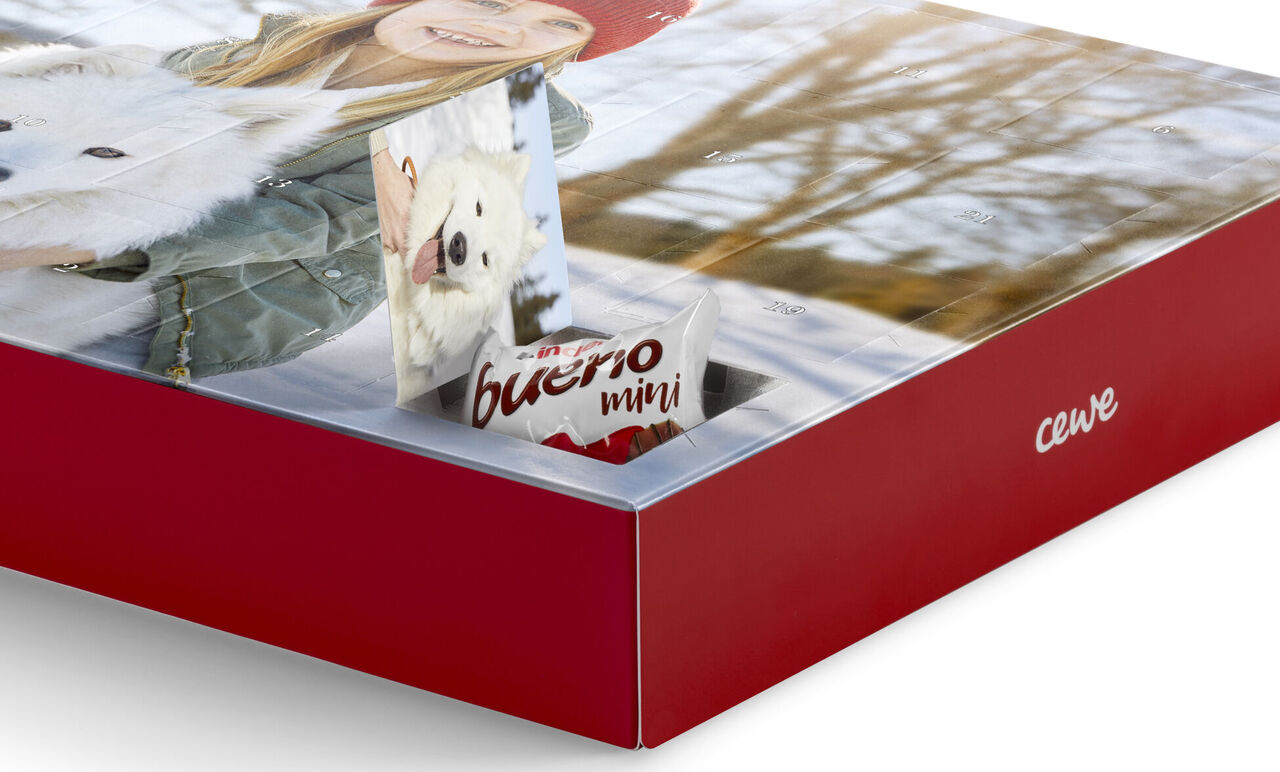 Una donna felice con un cane tiene un calendario fotografico CEWE e un pezzo di cioccolato su un tavolo.